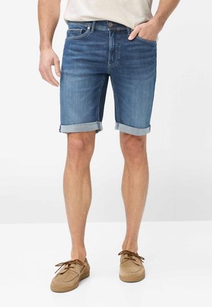 Mand iført medium blå opkoblede denimshorts, beige mokassin-sko og en hvid skjorte, med den ene hånd i lommen, stående mod hvid baggrund.