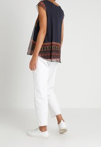 Kvinde iført en ærmeløs navyblå top med røde mønstre, hvide korte jeans og hvide sneakers, stående mod en ensartet lys baggrund.