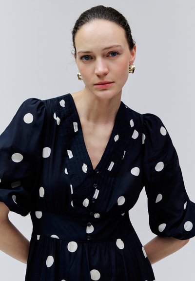 Kobieta o jasnej karnacji i niebieskich oczach ubrana w czarną sukienkę w białe kropki oraz złote kolczyki, stojąca na tle jednolitego jasnoszarego tła.