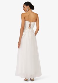 Robe blanche sans bretelles avec un corsage en dentelle florale, des lacets dans le dos et une jupe en tulle fluide. Présente des textures délicates et une coupe ajustée.