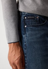 Close-up of a hand resting beside dark blue denim jeans pocket with visible brand label and gray long-sleeve sweater sleeve.