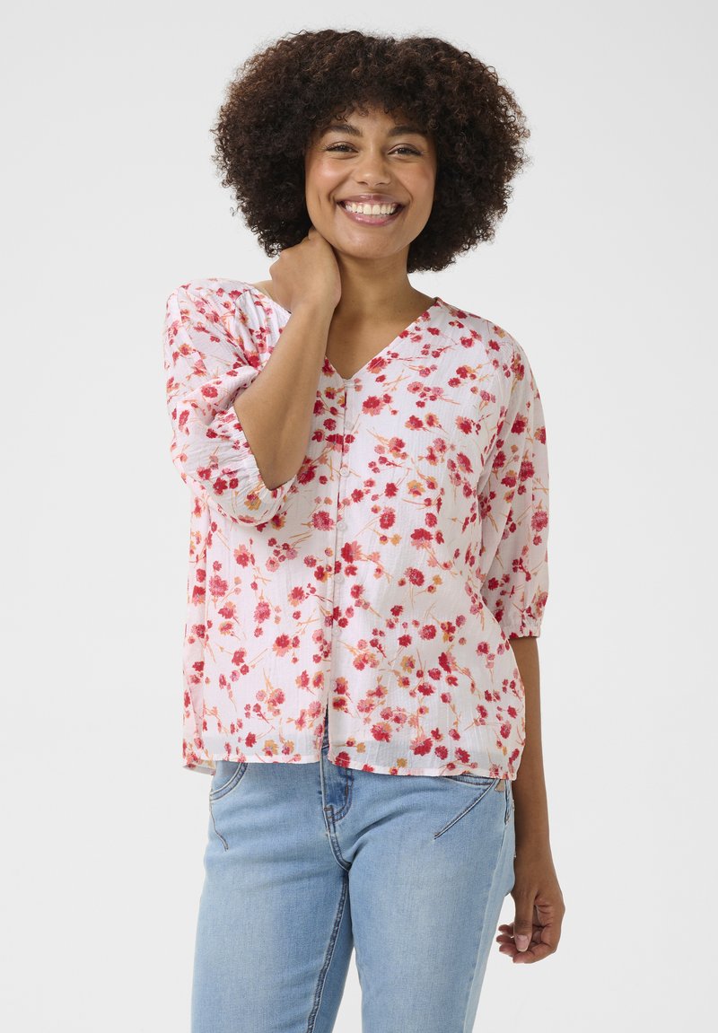 Blusa floreale rosa con maniche corte, scollo a V, vestibilità ampia, tessuto texture, piccoli fiori rossi, abbinata a jeans blu.