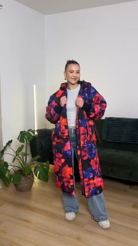 Žena v dlhom farebnom prešívanom kabáte s kvetinovým vzorom, sivom krátkom topu, modrých džínsach a bielych teniskách stojí v obývacej izbe s rastlinami a sedačkou.