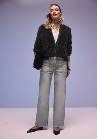 Jeune femme portant une veste noire texturée zippée, un chemisier blanc, un jean gris à jambes larges et des ballerines pointues noires, debout devant un fond dégradé uni.