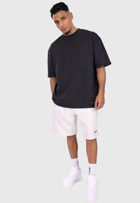 Sort oversized t-shirt med korte ærmer; parret med hvide shorts. Hvide sneakers og sokker med sorte striber fuldender outfittet. Simpelt design.