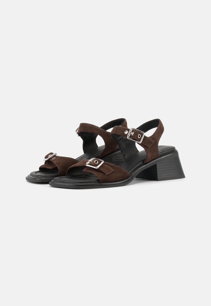 Braune Wildleder-Sandalen mit offenen Zehen, schwarzen klobigen Absätzen, Knöchelriemen und silbernen Schnallenverschlüssen vor weißem Hintergrund.