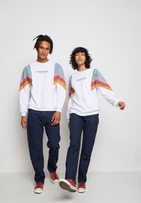 Hvid sweatshirt med multicolor skrå striber i lyseblå, pink og gul på skuldrene; båret med mørke jeans og røde sneakers.