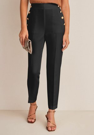 Femme portant un pantalon noir taille haute avec des boutons dorés sur les côtés, tenant une pochette bronze et portant des talons bronzes à brides.