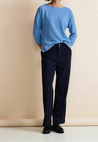 Ljusblå texturerad tröja med trekvartsärmar, i kombination med marinblå vida byxor med två framknappar. Accessoarer inkluderar svarta skor.