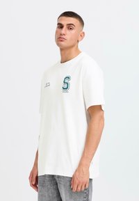 White cotton t-shirt with short sleeves, featuring green embroidered "S" logo on the left chest, worn with grey denim jeans.