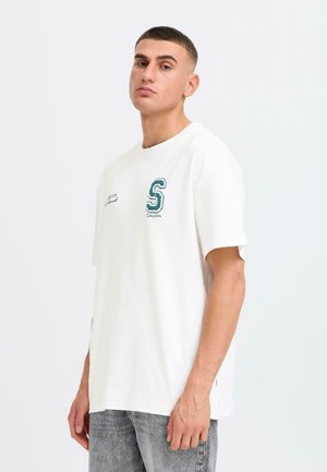 T-shirt en coton blanc à manches courtes, arborant un logo "S" brodé en vert sur la poitrine gauche, porté avec un jean en denim gris.
