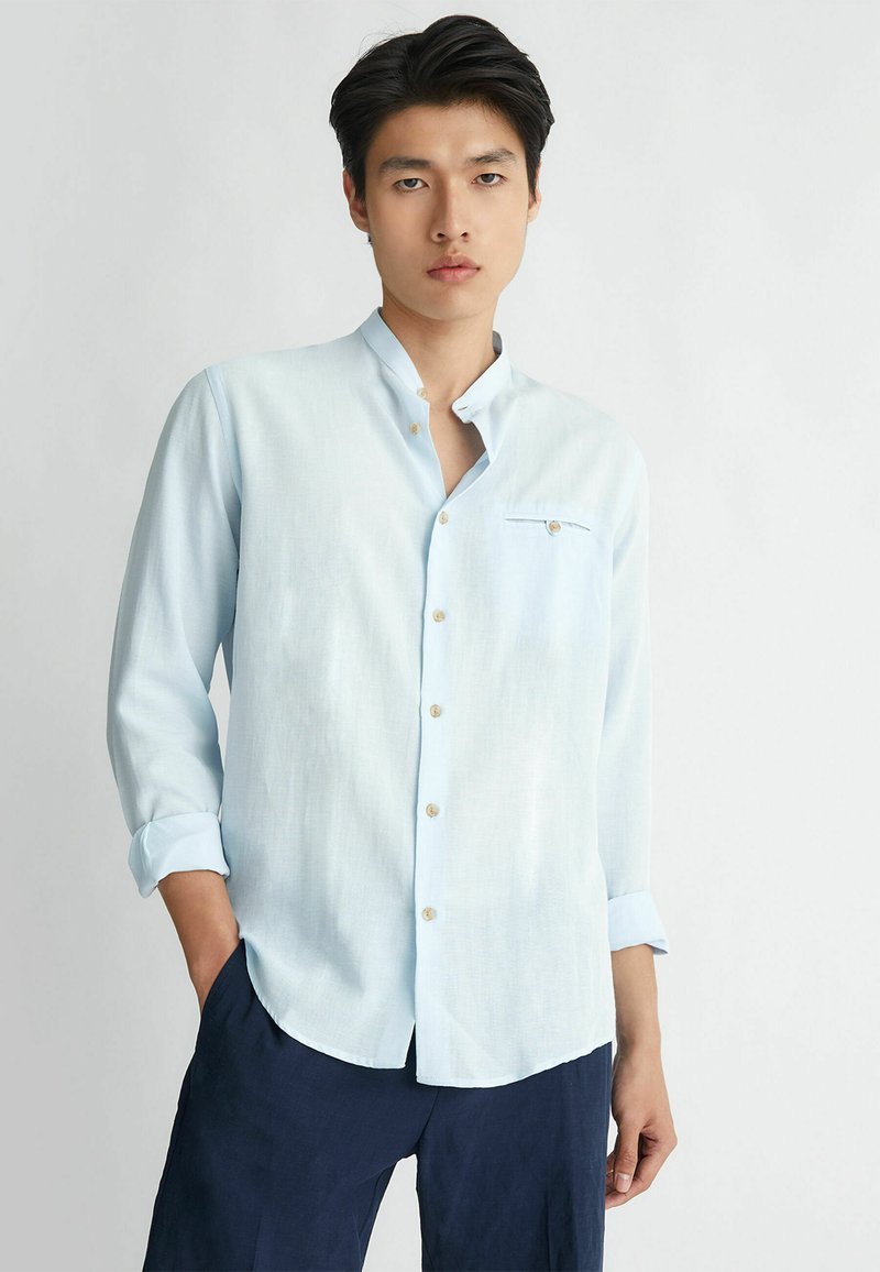 Camicia button-up blu chiaro con colletto alla coreana, maniche lunghe e taschino sul petto. Realizzata in un tessuto testurizzato, presenta bottoni dorati.