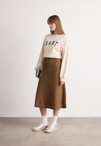 Kvinna som står inomhus iförd en krämfärgad sweatshirt med "GANT"-logotyp, brun midiklänning, vita strumpor, silverfärgade sneakers och håller en liten bok.