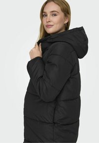 Chaqueta puffer negra con capucha, presenta un diseño acolchado y un corte relajado. La tela tiene una textura suave y detalles de costura visibles.