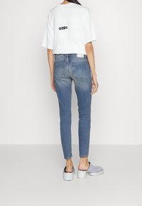 Camiseta blanca recortada con estampado negro "032c", combinada con jeans ajustados azules. El calzado incluye zapatillas gris oscuro sin cordones con tacones metálicos.