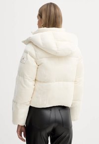 Giacca bianca gonfiabile con cappuccio, caratterizzata da sezioni trapuntate e un marchio sulla manica sinistra. Indossata sopra pantaloni in pelle nera.