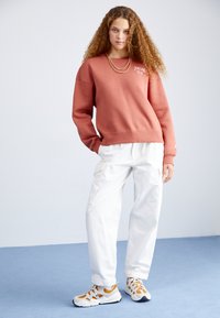 Sweatshirt en terre cuite avec logo blanc, associé à un pantalon cargo blanc ample. Les chaussures présentent des accents blancs et orange, le tout sur un fond neutre.
