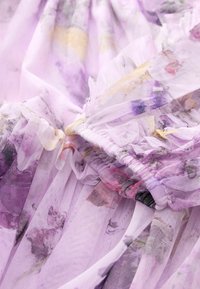 Lavendel-Tüllstoff mit floralen Mustern in rosa und gelben Farbtönen. Rüschendetails entlang der Kanten mit einer weichen, durchsichtigen Textur.