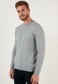 Maglione grigio strutturato con collo rotondo e polsini e orlo a costine. Il tessuto presenta un motivo a nido d'ape. Indossato con jeans scuri.