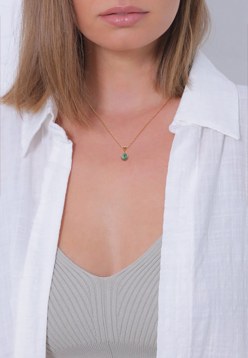 Gold chain necklace featuring a small pendant with a green stone, worn over a gray ribbed top, layered under a white shirt.