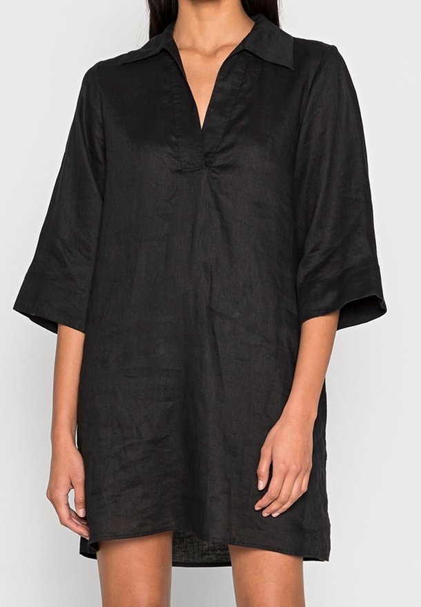 Femme portant une robe ample en lin noire avec des manches trois-quarts et un col en V, debout devant un fond blanc.
