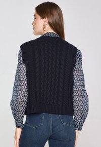 Gilet lavorato a maglia blu navy con un motivo a treccia testurizzato, design senza maniche, scollatura e orlo a coste, abbinato a una camicia a maniche lunghe con fantasia sotto.