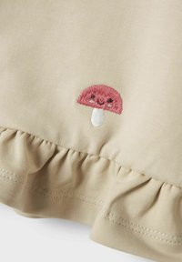 Vêtement en coton beige avec un ourlet froncé et un design de champignon brodé rose avec un visage souriant. Texture lisse, style décontracté.