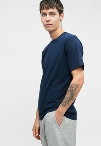 Camiseta de algodón azul marino con mangas cortas, cuello redondo y un ajuste relajado, combinada con pantalones de chándal grises. Presenta un tatuaje visible en el brazo.