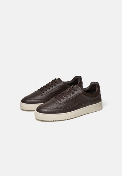 Paar donkerbruine leren sneakers met witte zolen en bruine veters op een lichtgrijze achtergrond.