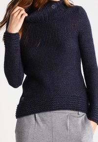 Navy-blauer Strickpullover mit einem Kragen und zwei Knöpfen, strukturiertem Stoff und einem bequemen Schnitt, kombiniert mit hellgrauen Hosen.