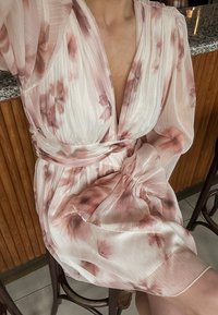 Roza cvetna obleka z zavihkom iz prosojnega blaga, globokim V-izrezom, širokimi rokavi in teksturiranim pasom, ki vsebuje nežne cvetlične vzorce po celotni obleki.