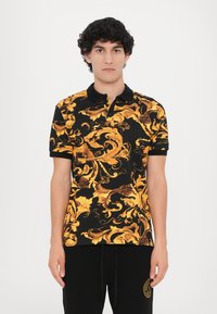 Camisa polo preta com um padrão floral dourado e castanho, mangas curtas e colarinho abotoado. Usada com calças de treino pretas.