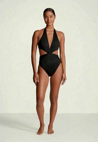 Mulher a pé descalço, vestindo um fato de banho preto de uma peça com decote halter e laterais recortadas, contra um fundo simples.