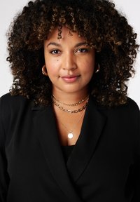 Zwarte blazer met een diepe V-hals, gelaagde gouden kettingen en grote gouden creolen. Krullend haar met highlights, een egale huid en neutrale make-up.