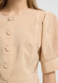 Ferskenfarvet bluse med rund halsudskæring, puffede ærmer og dekorative knapper. Tekstiler med lette gyldne prikker gennem hele stoffet.