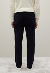 Marineblauwe ribfluwelen broek met een taps toelopende pasvorm, voorzien van achterzakken en een subtiele structuur. Draagt een lichtcrème trui en witte sneakers.