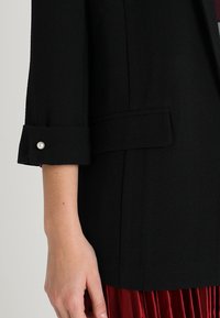 Ženská ruka blízko rukávu černého saka s perlou, pod ním je viditelná červená plisovaná sukně.