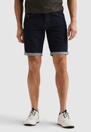 Man draagt donkere, opgerolde denimshorts, witte sneakers en een donkergroene shirt, staande tegen een effen achtergrond.