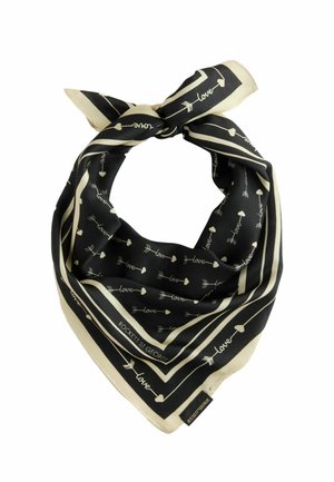 Frau lächelt mit schwarzen Haaren, trägt ein weißes kurzärmeliges Oberteil, einen schwarzen gemusterten Schalsgürtel, schwarze Hose, goldene Halskette und hält eine schwarze Handtasche.