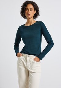 Frau mit lockigem Haar, die ein dunkelblaues Langarmshirt und cremefarbene Hosen trägt, steht mit den Händen in den Taschen vor einem schlichten Hintergrund.