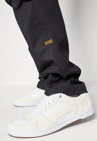Weiße Sneakers mit perforierten Akzenten und grünen Details, kombiniert mit schwarzen Hosen mit einem gelben G-Star-Label. Glatte Textur und klassisches Design.