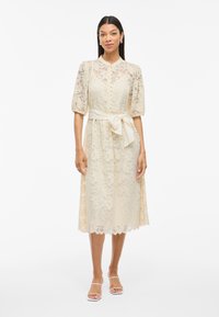 Lace midi klänning i krämfärg med korta puffärmar, knappstängning framtill och ett matchande midjebälte. Utsmyckad fållkant, mjuk textur.