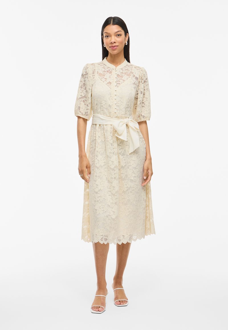 Lace midi klänning i krämfärg med korta puffärmar, knappstängning framtill och ett matchande midjebälte. Utsmyckad fållkant, mjuk textur.