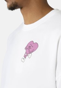 Fehér pamut póló, rózsaszín szív karakter grafika látható rajta, amelynek rajzfilmszerű karjai és lábai vannak, fehér cipőt visel, a mellkason elhelyezve.