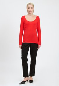 Rotes Langarmoberteil mit Rundhalsausschnitt, kombiniert mit schwarzen Hosen. Das Oberteil hat eine glatte Textur und eine figurbetonte Silhouette.