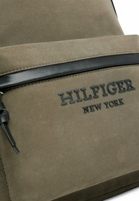Zaino color khaki con un tessuto texturizzato, dotato di una tasca zip nera in pelle e del logo in rilievo "HILFIGER NEW YORK".