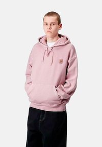 Sudadera con capucha de color rosa claro, con un bolsillo frontal y puños acanalados. Presenta un pequeño parche con el logo en la zona del pecho. Se lleva con pantalones negros.