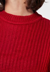 Primo piano di una persona che indossa un maglione rosso a coste con scollo arrotondato, che mostra il collo e parte dei capelli castani.