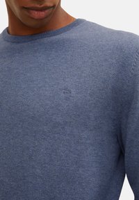 Blå stickad tröja i mjukt material, med rund halsringning och subtil logotypdetalj på bröstet. Slät textur och åtsittande design.