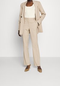 Costume beige à rayures avec une veste cintrée et un pantalon droit, associé à un haut côtelé crème et des talons dorés à motif serpent.